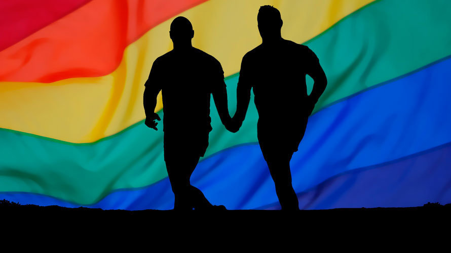 Una pareja camina de la mano con la bandera arcoiris por detrás. ARCHIVO.