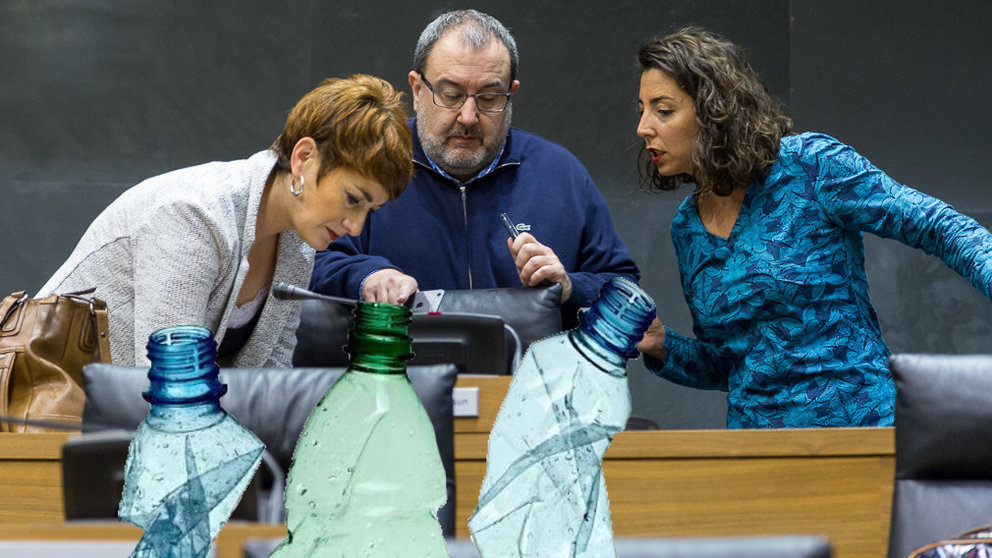 Bakartxo Ruiz, José Miguel Nuin y Laura Pérez conversan durante el pleno del Parlamento foral que ha tomado en consideración la nueva ley foral de residuos, junto a una imagen de unas botellas de agua embotellada NAVARRACOM